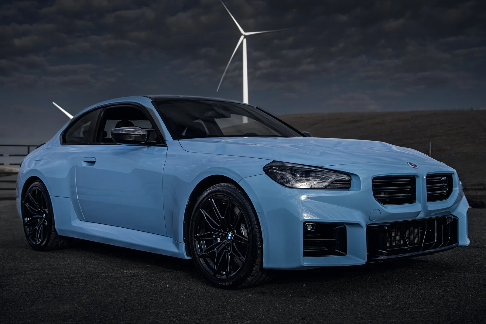 Blue 2023 BMW M2 under a dark sky with a wind turbine behind, front three-quarter view