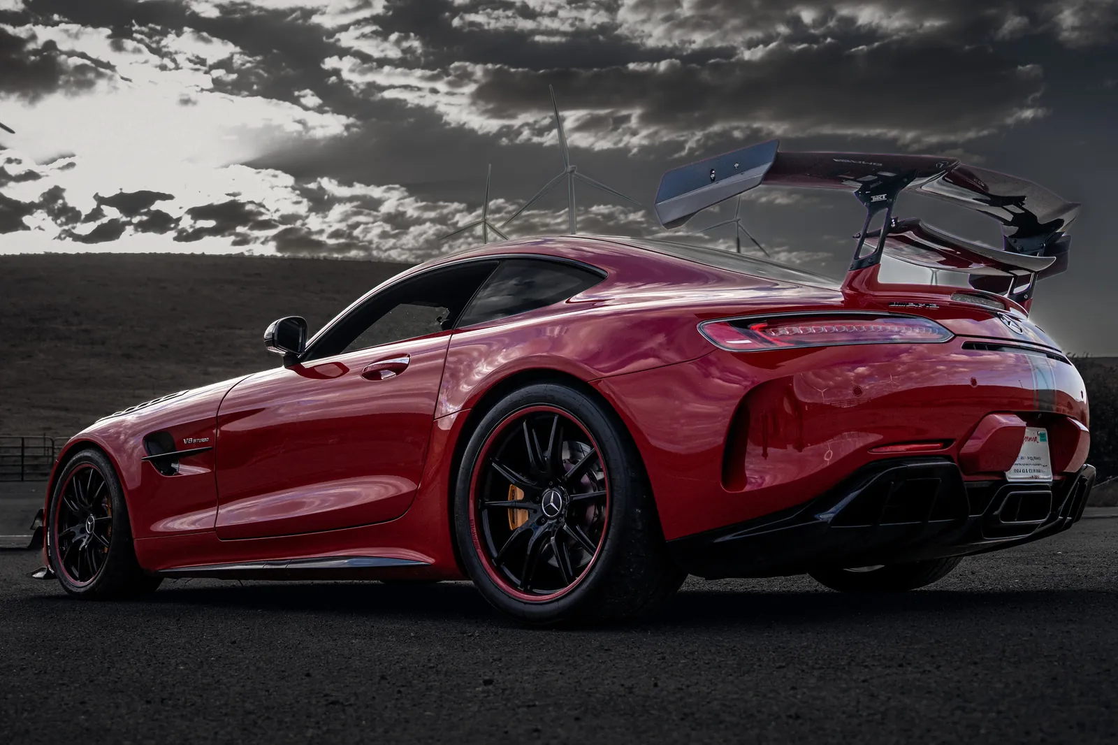 Red Mercedes-AMG GT R with a large rear wing, rear three-quarter at a low angle, sun and wind turbines behind