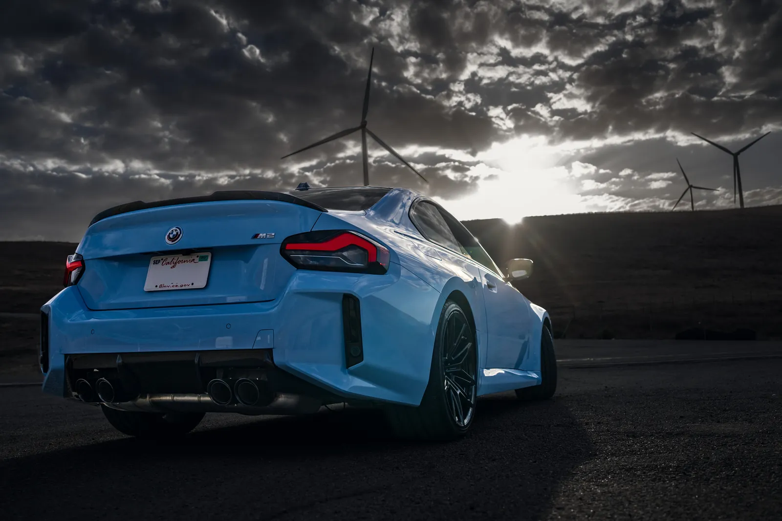 BMW M2 rear three-quarter with the sun breaking through clouds behind wind turbines
